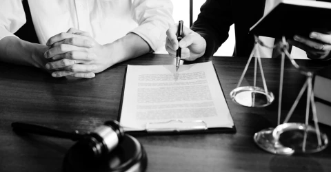 Two people signing a legal document across a desk beside a gavel and a set of scales.